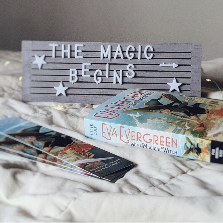Image of sign that reads 'The Magic Begins' and a copy of 'Eva Evergreen, Semi-Magical Witch' on a bed with some bookmarks to match the book cover