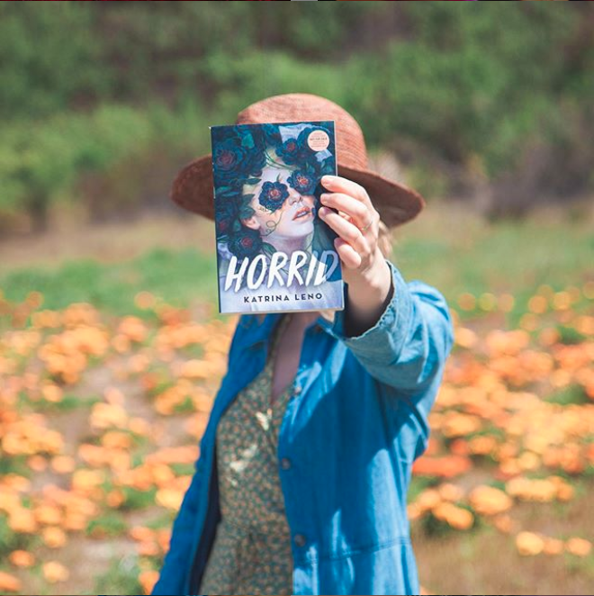 Katrina Leno holding up the book 'Horrid' by Katrina Leno, in a field during the day