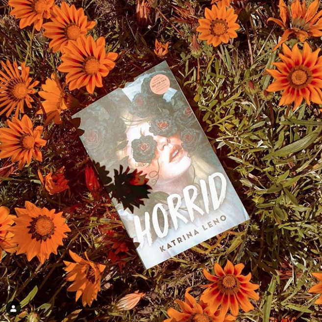 Picture of the book 'Horrid' by Katrina Leno in the grass, surrounded by orange flowers