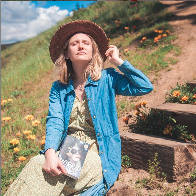 Katrina Leno wearing straw hat and dress, sitting on the side of the mountain, looking into the sun while holding the book 'Horrid' by Katrina Leno